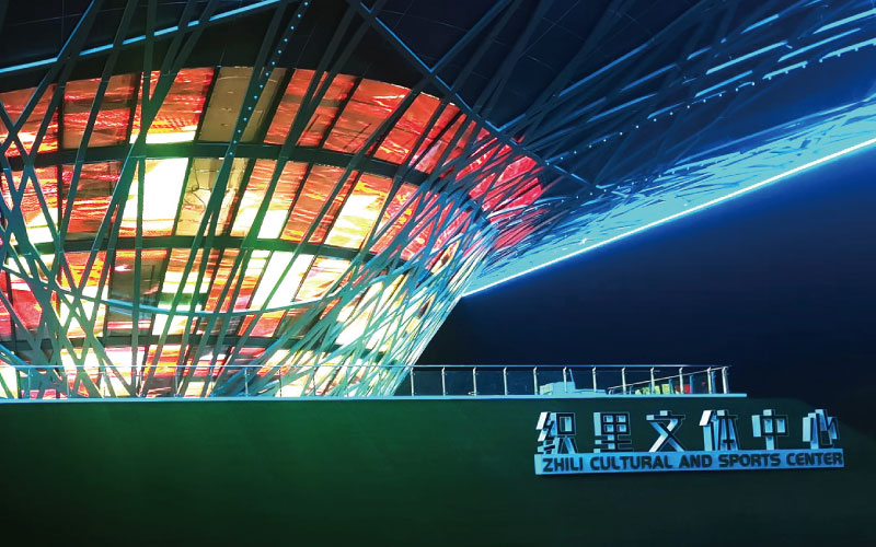 位于浙江湖州的織里文體中心，是一座大型異形鋼架結(jié)構(gòu)建筑。三思為其定制近1000平米透明屏，運用高清LED透明屏及控制系統(tǒng)，將其打造成了一幅巨型建筑媒體立面。
